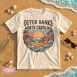 May include: Cream-colored t-shirt featuring a beach scene with the text "Outer Banks North Carolina." The graphic includes palm trees, a lifeguard stand, waves, and a sunset. The shirt is displayed on a sandy surface with seashells and sunglasses.
