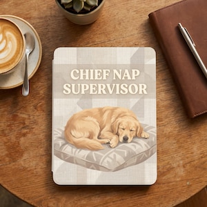 Op de afbeelding: Een notitieboek met de tekst "CHIEF NAP SUPERVISOR" en een illustratie van een slapende golden retriever op een kussen. Het notitieboek ligt op een houten tafel, naast een kop koffie en een vetplant.
