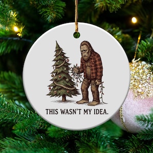 May include: A round white Christmas ornament featuring a Bigfoot character in a red and brown plaid shirt, holding Christmas lights next to a decorated tree. The text "THIS WASN'T MY IDEA." is printed below the image.