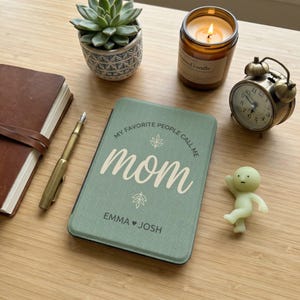 May include: A green journal with the text "My favorite people call me mom" and the names "Emma" and "Josh". The journal is next to a brown leather-bound notebook, a gold pen, a succulent plant, a lit candle, a small alarm clock, and a small figurine.
