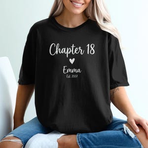 May include: Black t-shirt with the text "Chapter 18" in a script font, a heart symbol, the name "Emma", and the year "Est. 2007". The shirt is a casual style, suitable for everyday wear.