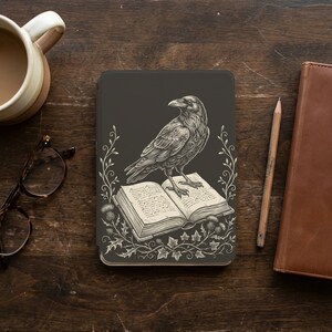 Puede incluir: Una funda de tableta gris oscuro con una ilustración detallada de un cuervo posado sobre un libro abierto. El libro está rodeado de elementos florales y de cardos. Un lápiz, gafas y una taza están sobre la superficie de madera.