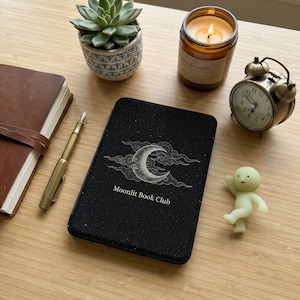以下が含まれることがあります： 木製の表面に、三日月模様と「Moonlit Book Club」の文字が入った黒いジャーナル。 ろうそく、小さな多肉植物、ヴィンテージの目覚まし時計もあります。 金色のペンと小さな置物が近くにあります。