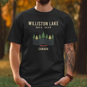 Puede incluir: Camiseta negra con un gráfico del lago Williston, Canadá. El diseño incluye las coordenadas del lago, una fila de árboles verdes y amarillos, líneas onduladas y una canoa roja. La palabra "CANADA" está impresa debajo.