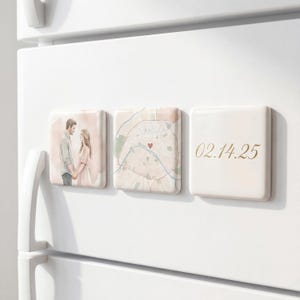 May include: Three square magnets on a white refrigerator door. The first magnet features a watercolor illustration of a couple holding hands. The second magnet displays a map with a heart. The third magnet has the date "02.14.25" in gold script.