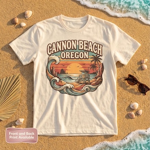 Peut inclure: T-shirt crème avec un motif de Cannon Beach, Oregon. Le design comprend un coucher de soleil, des vagues océaniques, une tour de sauveteur et des palmiers. Le texte "CANNON BEACH OREGON" est en arc. Des lunettes de soleil et des coquillages sont sur le sable.
