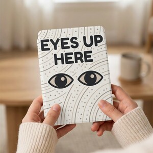 May include: A white tablet case with the text "EYES UP HERE" above a pair of stylized eyes. The case has a beige and gray pattern. The case is held in front of a blurred background.