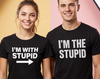 Może przedstawiać: Dwie osoby w pasujących czarnych koszulkach z białym tekstem. Jedna koszulka mówi "I'M WITH STUPID" ze strzałką w prawo. Druga koszulka mówi "I'M THE STUPID". Tło jest podzielone na żółty i różowy.