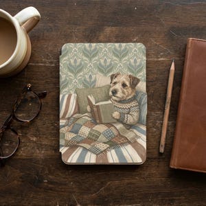 Puede incluir: Una funda decorativa para tableta con una ilustración de un perro leyendo un libro. El perro lleva un suéter y está acostado en una cama con una colcha de retazos y almohadas. El fondo tiene un estampado floral.