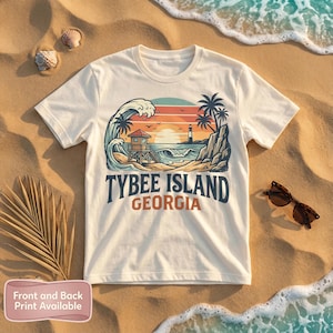 May include: Cream-colored t-shirt featuring a beach scene with a lifeguard tower, lighthouse, and palm trees. The text "TYBEE ISLAND GEORGIA" is printed below the graphic. Sunglasses rest on the sand.