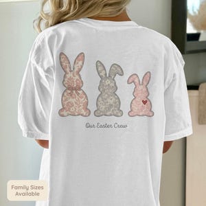 Puede incluir: Camiseta blanca con tres siluetas de conejos en patrones florales rosas y grises. Los conejos están dispuestos en fila, con el texto "Our Easter Crew" debajo. Un pequeño corazón está en el conejo de la derecha.