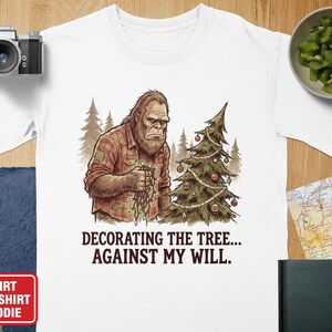 May include: White t-shirt with a graphic of a Bigfoot figure decorating a Christmas tree. The Bigfoot is wearing a plaid shirt and holding Christmas lights. Text on the shirt reads, "Decorating the tree... against my will."