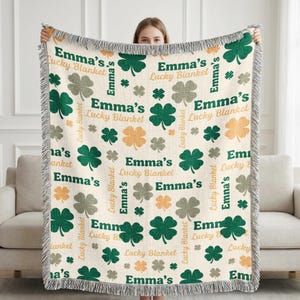 以下が含まれることがあります： クリーム色の織りブランケット。緑、グレー、オレンジのクローバーの繰り返し模様と「Emma's Lucky Blanket」の文字が特徴です。 ブランケットはフリンジ加工が施されており、人が持っています。