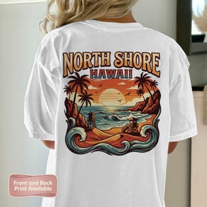 Puede incluir: Camiseta blanca con un diseño gráfico colorido de una escena de playa hawaiana y el texto "NORTH SHORE HAWAII". El diseño incluye palmeras, olas del mar y una puesta de sol.