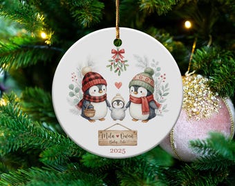 May include: A round Christmas ornament featuring a family of penguins wearing winter hats and scarves. The ornament has a wooden sign with the names "Mila & Owen" and "Baby Arlo" with the year 2025. The ornament is hanging on a Christmas tree.
