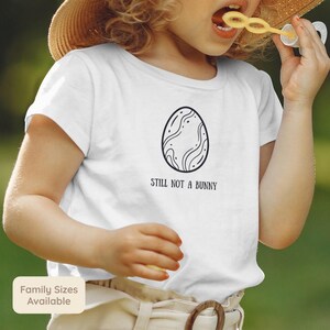 May include: A white t-shirt with a black egg design and the words "STILL NOT A BUNNY". A child is wearing the t-shirt and blowing bubbles outdoors.