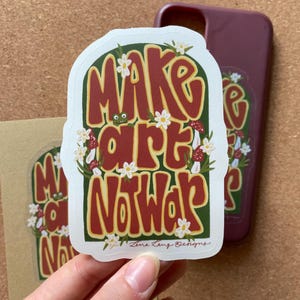 Könnte beinhalten: Ein weißer Aufkleber mit dem Text "MAKE ART NOT WAR" in einer Retro-Schrift, umgeben von Blumen und Pilzen. Eine passende Handyhülle und ein Aufkleber sind im Hintergrund zu sehen. Der Aufkleber wird von einer Hand gehalten.