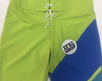 May include: Lime green swim trunks with a blue and teal diagonal stripe pattern. The trunks have a white drawstring and a white circle patch with the text "IFBB SHOP" in black.