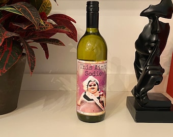 May include: A white wine bottle with a label that says "This Ain't Sodie". The label features a cartoon illustration of a woman wearing a pink dress and holding a glass of wine.