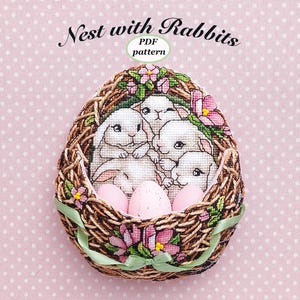 May include: A cross-stitch pattern for a nest with four baby rabbits and three Easter eggs. The nest is made of woven twigs and has pink flowers and green leaves. The pattern is titled "Nest with Rabbits" and is available as a PDF download.
