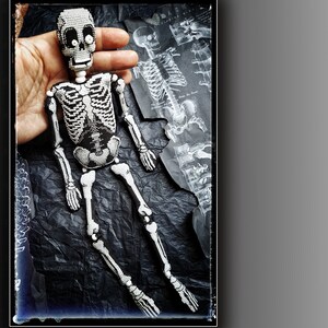 May include: A beaded skeleton doll with white beads and black thread. The doll is posed sitting with its legs crossed. The doll is on a black background.