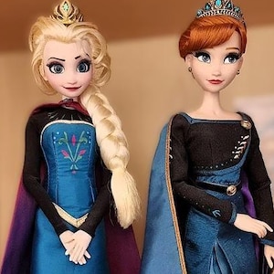 以下が含まれることがあります： アニメのキャラクターの人形が2体。1体は金髪を三つ編みにし、青いドレスと紫のマントを着ています。もう1体は赤毛で、青いドレスと青と紫のマントを着ています。どちらの人形も王冠をかぶっています。