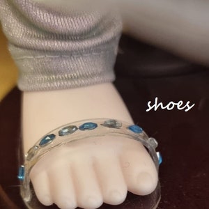 May include: Close-up of a doll's foot wearing a clear sandal adorned with blue gemstones. The sandal's strap curves over the toes. The doll's leg is covered in a gray fabric. The word "shoes" is written in cursive.