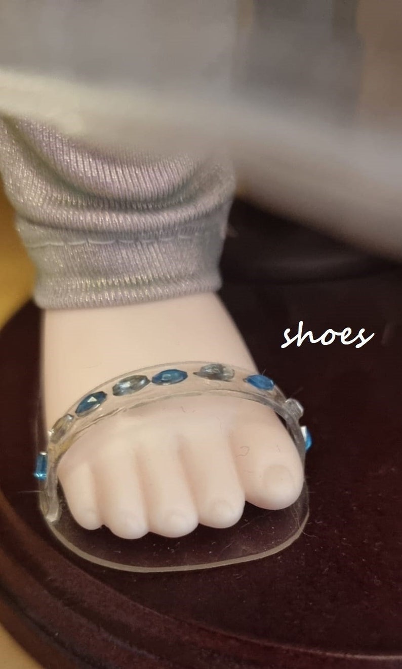 May include: Close-up of a doll's foot wearing a clear sandal adorned with blue gemstones. The sandal's strap curves over the toes. The doll's leg is covered in a gray fabric. The word "shoes" is written in cursive.
