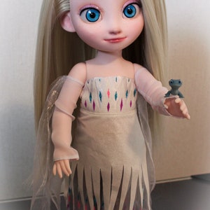 May include: A custom doll of a character with long blonde hair and blue eyes, wearing a white dress with a decorative pattern and a sheer overlay. The doll is holding a small blue figurine. The text "SahoryCustom" is visible.