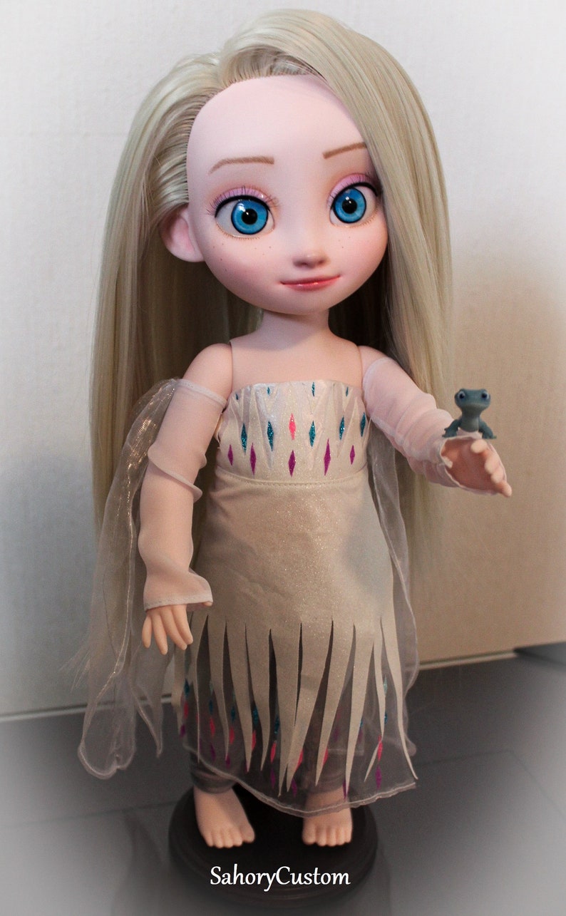 May include: A custom doll of a character with long blonde hair and blue eyes, wearing a white dress with a decorative pattern and a sheer overlay. The doll is holding a small blue figurine. The text "SahoryCustom" is visible.