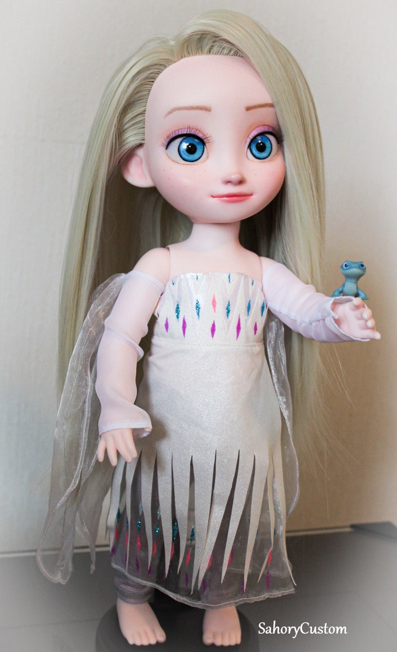 May include: A custom doll of a character with long, light blonde hair and blue eyes. The doll wears a white dress with a decorative pattern and a sheer cape. A small blue frog figurine is held in the doll's hand.