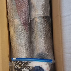 以下が含まれることがあります： 段ボール箱の中に、気泡緩衝材で包まれた商品がいくつか入っています。「Toppits ZIPPER」の文字が入ったジッパー付きバッグが見え、他の商品も入っています。バッグには青と白の雪の結晶のデザインが施されています。