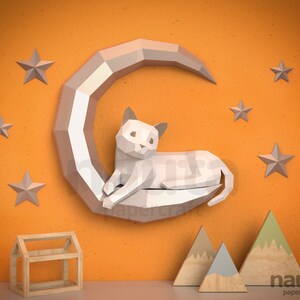 May include: A white papercraft cat rests on a crescent moon, surrounded by grey stars. The cat and moon are mounted on an orange wall. Wooden house and mountain decorations are in the foreground. The image is labelled "nauta papercraft."