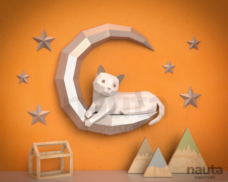 May include: A white papercraft cat rests on a crescent moon, surrounded by grey stars. The cat and moon are mounted on an orange wall. Wooden house and mountain decorations are in the foreground. The image is labelled "nauta papercraft."
