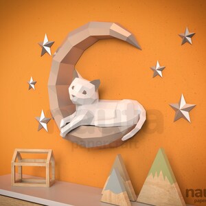 May include: A papercraft cat on a crescent moon, with stars, against an orange wall. The cat and moon are light brown and white. Additional wooden house and mountain papercrafts are on a shelf below. The word "nauta papercraft" is in the bottom right corner.