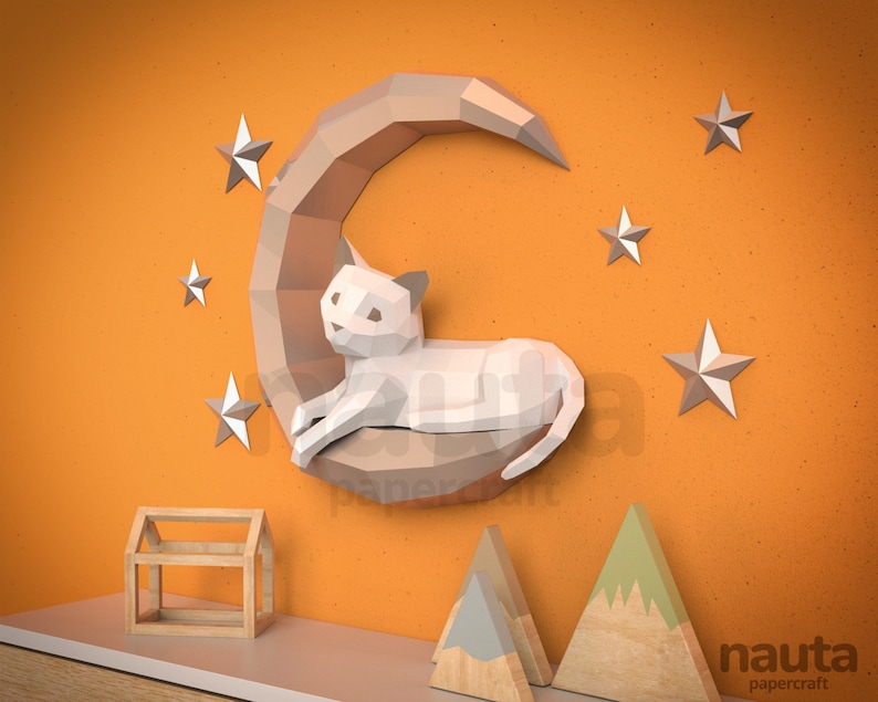 May include: A papercraft cat on a crescent moon, with stars, against an orange wall. The cat and moon are light brown and white. Additional wooden house and mountain papercrafts are on a shelf below. The word "nauta papercraft" is in the bottom right corner.