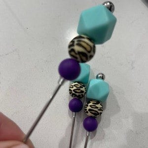 以下が含まれることがあります： さまざまなビーズが付いた、3つの装飾用串のセット。各串には、紫色の球体、ヒョウ柄の球体、水色の立方体が付いています。串は銀色のビーズで飾られています。