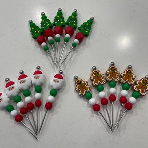 Peut inclure: Ensemble de six piques à nourriture sur le thème de Noël avec des perles vertes, rouges et blanches. Les piques représentent le Père Noël, des hommes en pain d'épices et des sapins de Noël.
