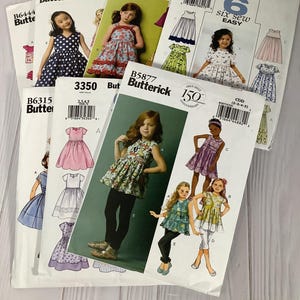 Może przedstawiać: Kolekcja wykrojów krawieckich Butterick na sukienki dla dziewczynek. Wzory przedstawiają ilustracje gotowych ubrań, o różnych stylach i wzorach. Okładki pokazują numery wzorów i logo Butterick.