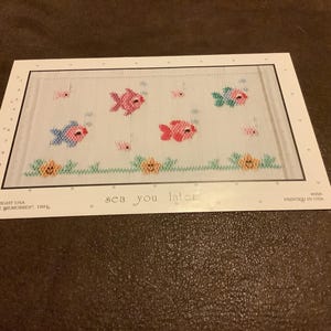 May include: A cross-stitch pattern featuring colorful fish and bubbles. The design includes pink, blue, and green fish, with the text "sea you later". The pattern is framed with a dotted border and a floral border at the bottom.