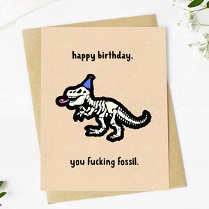 May include: A birthday card with a tan background and a brown envelope. The card features a black and white dinosaur skeleton wearing a party hat and sticking out its tongue. The text reads "happy birthday, you fucking fossil."