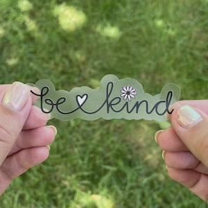 May include: A clear sticker with the text "be kind" in black script with a small heart and a pink flower graphic.