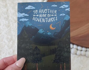 May include: A greeting card with a night scene illustration. The card features a dark blue sky with white clouds, stars, and a crescent moon. The text "TO ANOTHER YEAR OF ADVENTURES" is written across the top. The bottom of the card shows a forest and mountains.