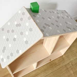 Puede incluir: Estantería de madera con forma de casa con un techo con estampado de escamas grises y blancas. La estantería tiene varios compartimentos y un bloque verde en la parte superior. La estantería es de madera clara.