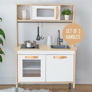 May include: White wooden play kitchen with a microwave, sink, stovetop, and two cabinets. The cabinets have leather handles. The kitchen has a wooden shelf above the microwave and a metal bar for hanging utensils. The text "SET OF 3 HANDLES" is on a brown circle.