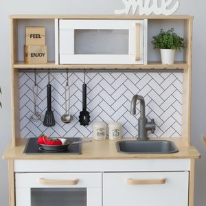 May include: A wooden toy kitchen with a white microwave, sink, and stove. It includes toy utensils, a frying pan with a tomato, and salt and pepper shakers. The backsplash has a black and white herringbone pattern. The word "Smile" is written above.