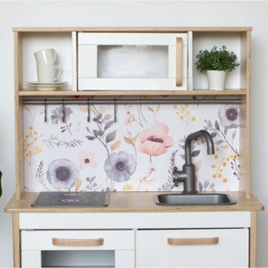 May include: A wooden toy kitchen with a white microwave and cabinets, featuring a floral backsplash with pink and purple flowers. Includes a sink, stovetop, and a small plant. The handles are light brown.