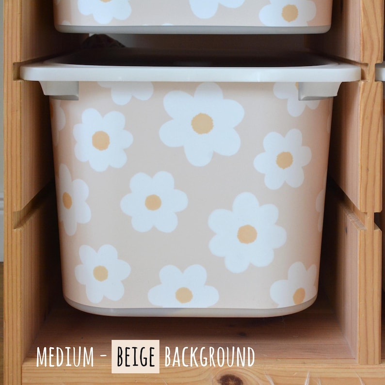 May include: Beige storage bin with a white daisy pattern and a yellow center. The bin has a white rim and is labeled "MEDIUM - BEIGE BACKGROUND". The bin is inside a wooden shelf.