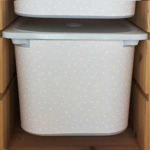 Puede incluir: Un contenedor de almacenamiento de plástico beige con un patrón de lunares blancos. El contenedor está dentro de una unidad de estantería de madera.