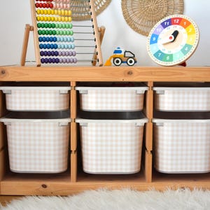 Pu&ograve; includere: Un mobiletto portagiochi in legno chiaro con tre colonne e sei contenitori in plastica bianca con motivo a quadretti beige e bianco. Il mobile ospita un pallottoliere colorato, un piccolo camion giocattolo e un orologio rotondo con numeri da 1 a 24.